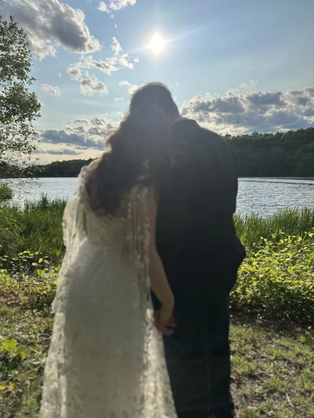 A couple standing together at the edge of a lake at sunset, seen from behind.
