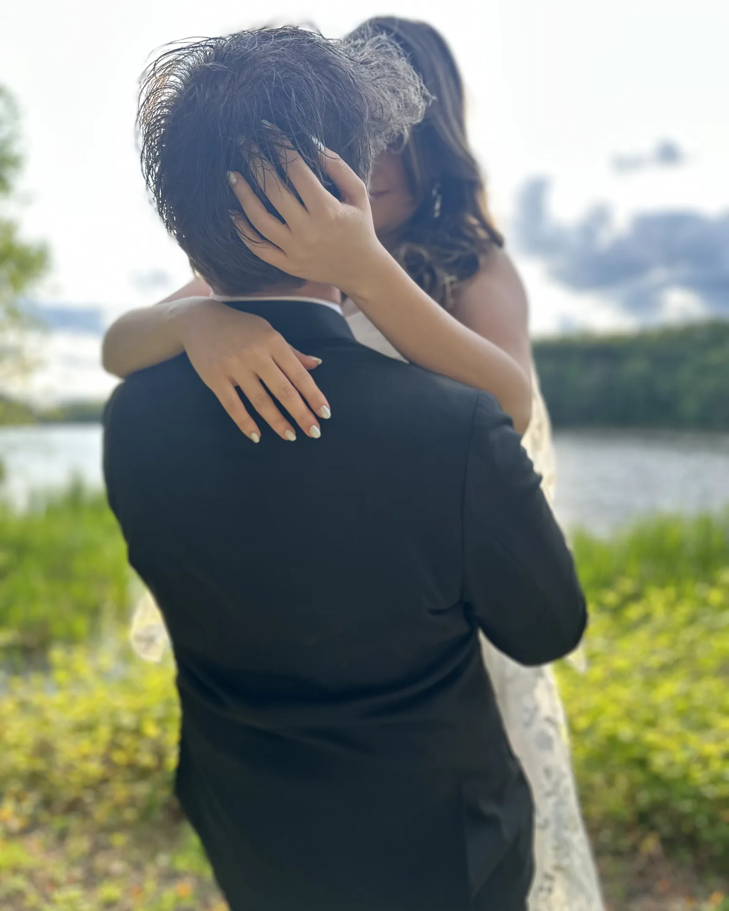 A couple holding each other close by a lake, her hands behind his head.