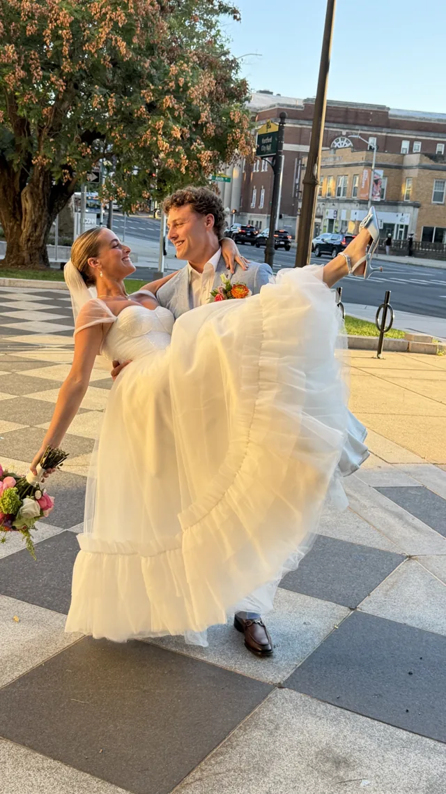 A groom carrying his bride along a city sidewalk, both of them laughing.