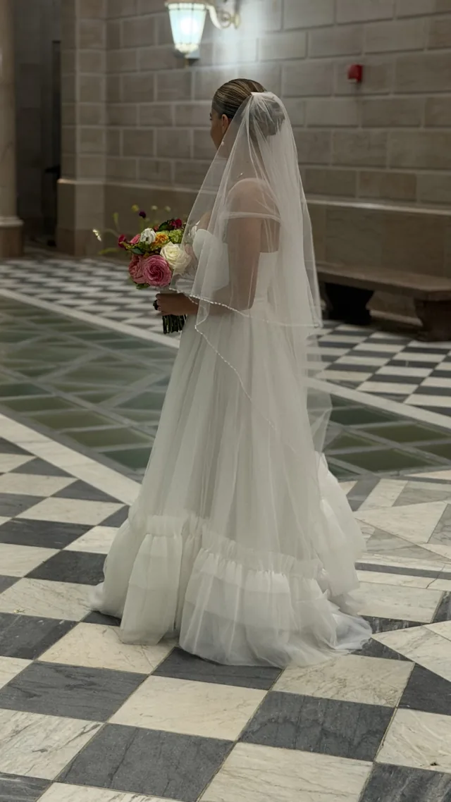 A bride from behind, holding her bouquet in a stone hallway with a checkered marble floor.